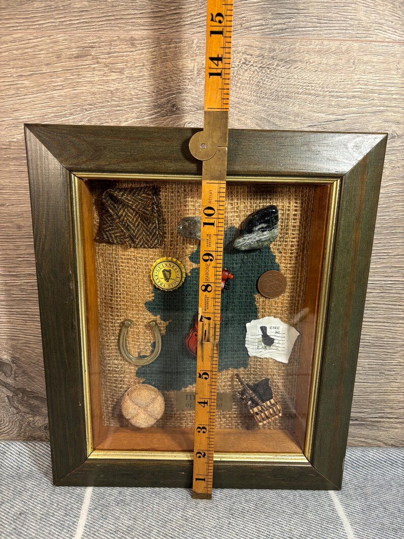 Puede incluir: Una caja de sombra enmarcada con un fondo de arpillera y varios art&iacute;culos de tem&aacute;tica irlandesa, incluyendo una herradura, un tr&eacute;bol, un peque&ntilde;o viol&iacute;n, una pelota de f&uacute;tbol, un trozo de tela de tweed, una peque&ntilde;a cesta, una moneda y un trozo de papel con el texto "C&iacute;rc", que en irland&eacute;s significa "c&iacute;rculo".