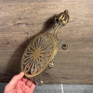 Puede incluir: Trivet de latón ornamentado con un diseño floral. El salvamanteles tiene un asa y patas decorativas. El centro presenta un patrón de rayos de sol. El latón tiene una pátina envejecida.