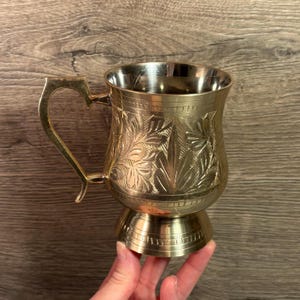 May include: A brass mug with an ornate floral pattern and a heart-shaped handle. The mug has a pedestal base and a polished interior. The mug is held in a hand against a wooden background.