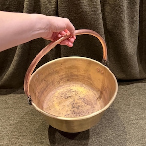 May include: A round brass bucket with a smooth, curved copper handle attached by metal rivets on each side. The interior of the brass basin shows signs of wear with dark, mottled discolouration across the bottom surface and along the inner walls.