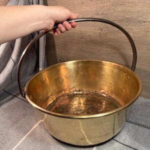May include: A large, antique brass pot with a worn finish and a sturdy iron handle. The pot has a rounded bottom and a wide, shallow opening.