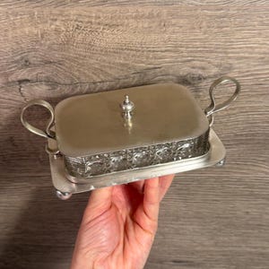 Op de afbeelding: Een rechthoekige botervloot met een heldere glazen basis met een gestructureerd ruitpatroon en een glad, zilverkleurig metalen deksel. Het deksel heeft een kleine centrale knop en de basis rust op een zilverkleurig metalen dienblad met twee handgrepen.