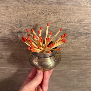 Op de afbeelding: Een kleine, antieke messing kom gevuld met houten lucifers. De lucifers hebben rode koppen en zijn waaiervormig gerangschikt. De kom heeft een geëtst ontwerp en wordt tegen een houten achtergrond gehouden.
