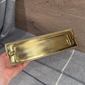 Könnte beinhalten: Eine rechteckige Briefkastenplatte aus Messing mit einem seilartigen Rand. Die Platte hat eine Klappe und einen Schlitz für Post. Das Messing hat eine polierte Oberfläche.
