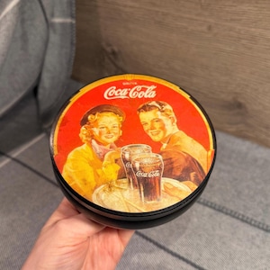 May include: Round black tin with a vintage Coca-Cola advertisement. The red lid features a smiling couple and glasses of Coca-Cola. The Coca-Cola logo is prominently displayed. The tin is held in a hand.