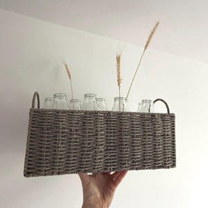 May include: A rectangular woven basket in shades of gray holds several clear glass bottles, each containing a single stem of dried wheat. The basket has two curved handles and is held up against a white background.