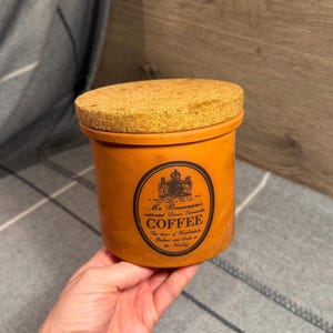 May include: A cylindrical terracotta coffee canister with a natural cork lid. The front features a black oval label with a crest and the text: Mr. Brannam's celebrated Devon Terracotta COFFEE, The choice of Housekeepers, Butlers and Cooks to the Nobility.