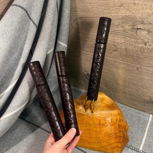 May include: Three dark brown leather-wrapped tools with a decorative stitched pattern. One tool has gold-colored prongs. The tools are displayed on a wooden base and against a gray and white plaid fabric.