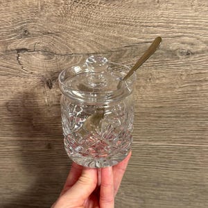 May include: A clear, cut-glass sugar bowl with a lid and a small spoon. The bowl has a diamond pattern and a round knob on the lid. The spoon is made of metal. The sugar bowl is held in a person's hand.