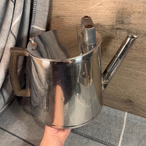 Puede incluir: Una tetera de metal plateado con un asa de madera y un pico. La tetera tiene una superficie brillante y reflectante.