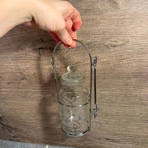 Pode incluir: Um frasco de vidro transparente com tampa e design floral gravado, preso em uma estrutura de metal prateado com uma alça e uma colher pequena. O frasco foi projetado para conter condimentos ou itens pequenos.