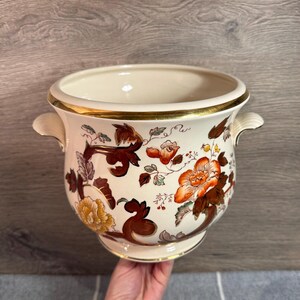 Puede incluir: Jardinera de cerámica color crema con borde y asas doradas. Presenta motivos florales en tonos naranja, marrón y amarillo. Un artículo decorativo para uso en interiores o exteriores.