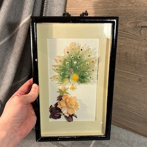 Può includere: Un'opera d'arte incorniciata con un pavone con la coda a ventaglio, realizzata con materiali iridescenti. Il pavone ha dettagli verdi e gialli ed è posizionato sopra un gruppo di fiori arancioni. La cornice è nera.