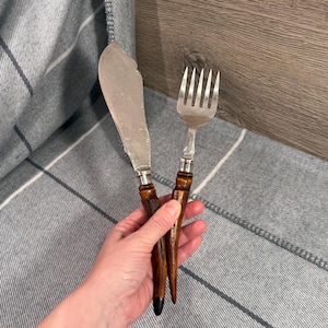 May include: A butter knife and fork set. The knife has a wide, slightly curved blade. Both utensils have silver metal tops and dark brown, pointed handles. The handles appear to be made of wood or a similar material. The set is displayed on a gray and white plaid surface.