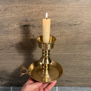 Puede incluir: Una vela encendida en un candelabro de latón. La vela es blanquecina y tiene una pequeña llama. El candelabro tiene una base redonda con un asa y un tallo decorativo.