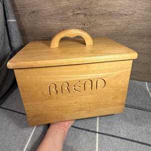 Könnte beinhalten: Eine hellfarbene Brotdose aus Holz mit einem gebogenen Griff auf dem Deckel. Das Wort "BREAD" ist in die Vorderseite der Box geschnitzt. Die rechteckige Box scheint aus massivem Holz zu sein.
