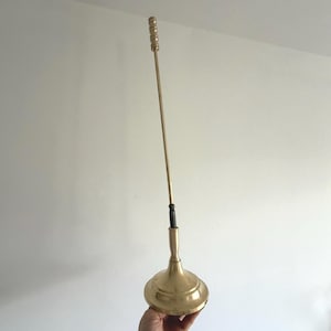 Könnte beinhalten: Ein Vintage-Flaschenverschluss aus Messing. Er hat einen langen, schlanken Stiel und eine dekorative Oberseite. Die Basis hat eine abgerundete, ausgestellte Form. Der Stiel ist mit kleinen, abgerundeten Akzenten versehen. Der Verschluss ist vor einem weißen Hintergrund.
