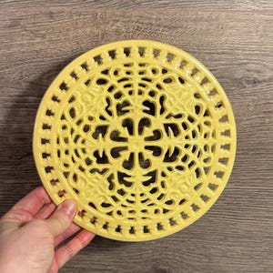 May include: A round, yellow ceramic trivet with an intricate, openwork design. The trivet features a central floral pattern surrounded by a decorative border. The trivet is held by a hand, showcasing its size and detail. This kitchenware item is designed to protect surfaces from heat.