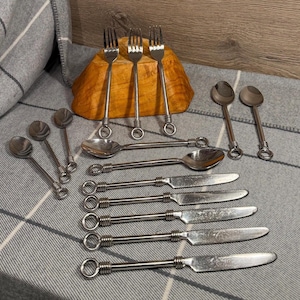 May include: A collection of stainless steel silverware, including forks, spoons, and knives, arranged on a gray and white patterned surface. The silverware has a decorative ring design on the handles. A wooden bowl is in the background.