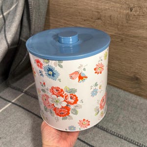 May include: A white metal canister with a blue lid and a floral design. The canister is decorated with a pattern of red, pink, blue, and yellow flowers with green leaves. The lid has a small, round handle.