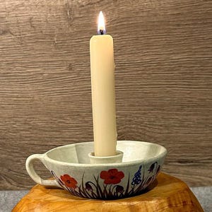Puede incluir: Un portavelas de cerámica blanca con un diseño floral con flores rojas y azules. El portavelas tiene un asa y una abertura central para una vela. Una vela blanca encendida se coloca en el portavelas.