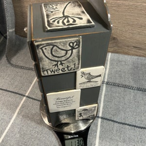 May include: A grey wooden box with a decorative top and a scale showing 1130 grams. The box has several small white tiles with black lettering and images of birds and flowers. The tiles have inspirational quotes, including "Tweet!" and "Beautiful things happen when you distance yourself from the negative."