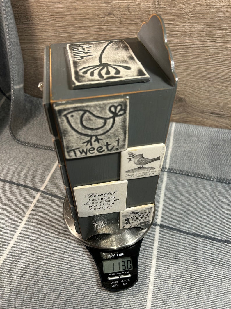May include: A grey wooden box with a decorative top and a scale showing 1130 grams. The box has several small white tiles with black lettering and images of birds and flowers. The tiles have inspirational quotes, including "Tweet!" and "Beautiful things happen when you distance yourself from the negative."