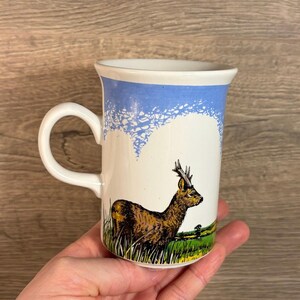 Puede incluir: Taza de cerámica blanca con una ilustración de ciervo. La taza presenta una banda azul en la parte superior y un asa curva. El ciervo es marrón y amarillo, de pie en un campo de hierba verde. Un pequeño árbol está en el fondo.