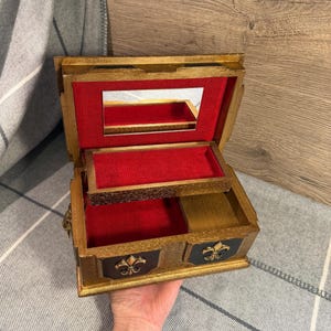 May include: Open wooden jewelry box with a gold finish, red velvet lining, and a small mirror. The box has a drawer with a black panel and gold fleur-de-lis design. The box is open, revealing the interior compartments.