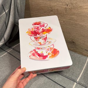 May include: A white rectangular tin featuring a watercolor illustration of three stacked teacups and saucers with red and orange floral designs. The tin has the text "M&S" on the bottom. The tin is resting on a gray and white plaid blanket.