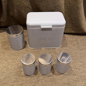 May include: A light grey metal bread bin with a hinged lid and the word BREAD printed on the front, accompanied by three matching cylindrical canisters labelled BISCUITS, TEA, SUGAR, and COFFEE, all featuring metallic silver-toned lids.