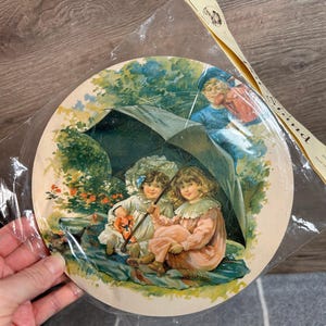Könnte beinhalten: Ein runder Vintage-Pappteller mit einer farbenfrohen Illustration von drei Kindern unter einem großen Regenschirm in einer Gartenlandschaft. Die Kinder tragen Hüte und Kleider. Der Regenschirm ist schwarz mit einem weißen Griff. Der Hintergrund ist eine grüne und blaue Landschaft mit Blumen und Bäumen.