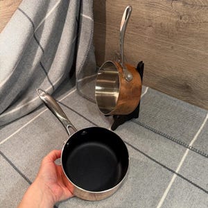 May include: Two stainless steel saucepans with long handles. One is copper-coloured and mounted on a black stand. The other has a black interior and is held by a hand. Both are on a grey and white plaid blanket.