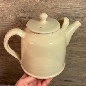 Puede incluir: Tetera de cerámica color crema con tapa y asa curva. La tetera tiene un pico y un pequeño pomo en la tapa. La tetera es de color beige claro y parece hecha a mano. La tetera está diseñada para preparar té.