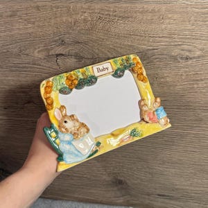 May include: A yellow ceramic baby photo frame featuring bunny rabbit figures and the word "Baby". The frame is rectangular with a white space for a photo. The frame is decorated with carrots and other foliage.