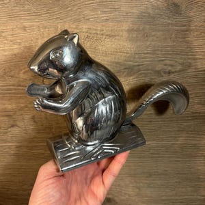 May include: A silver squirrel nutcracker with a detailed design. The squirrel is sitting on a log and has its tail curled around its body. The nutcracker is made of metal and has a shiny finish.