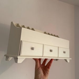 Puede incluir: Organizador de pared de madera blanquecina con tres cajones pequeños y una sección superior con múltiples ranuras. El organizador está sostenido por una mano contra una pared blanca.