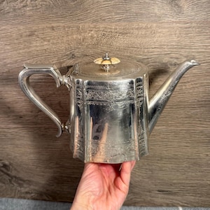 May include: A silver teapot with an elaborate design. The teapot features a curved handle, a spout, and a lid with a decorative knob. The body of the teapot has a faceted shape and is decorated with etched floral patterns.