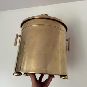 Könnte beinhalten: Ein großer, zylindrischer Messingbehälter mit Deckel, zwei Holzgriffen und drei abgerundeten Füßen. Der Behälter hat eine polierte Oberfläche und eine leicht konische Form. Der Deckel hat einen kleinen, runden Knopf. Der Behälter wird von einer Hand gehalten.
