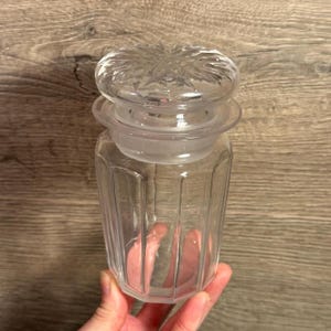May include: A clear glass jar with a decorative lid. The jar has vertical ridges and a round, frosted glass lid with a starburst design. The jar is held in a person's hand.