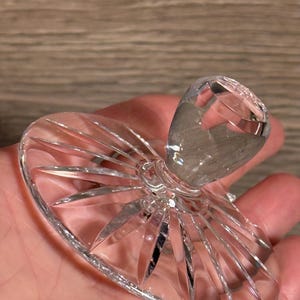 Op de afbeelding: Een heldere, facetgeslepen kristallen flesstopper. De stopper heeft een afgeronde bovenkant en een uitlopende basis met een zonnestraalpatroon. Het kristal reflecteert licht, waardoor een sprankelend effect ontstaat. Het ontwerp is elegant en decoratief.