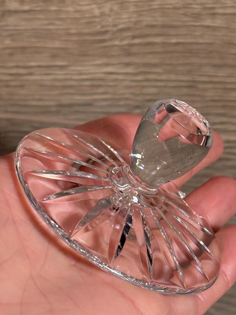 Op de afbeelding: Een heldere, facetgeslepen kristallen flesstopper. De stopper heeft een afgeronde bovenkant en een uitlopende basis met een zonnestraalpatroon. Het kristal reflecteert licht, waardoor een sprankelend effect ontstaat. Het ontwerp is elegant en decoratief.