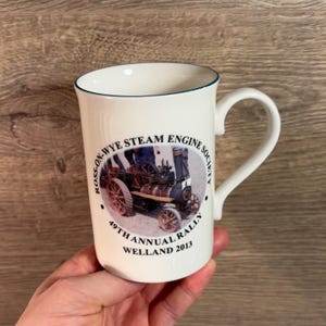 May include: White ceramic mug with a green rim and a black and white image of a steam engine. The text on the mug reads "Ross-on-Wye Steam Engine Society 49th Annual Rally Welland 2013".