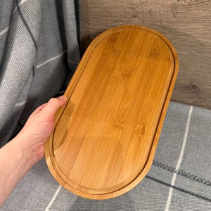 May include: An oval-shaped bamboo serving tray with a smooth, light brown wood grain finish. The tray features a raised outer rim and a flat, recessed center surface, showcasing the natural texture and color variations of the bamboo material.