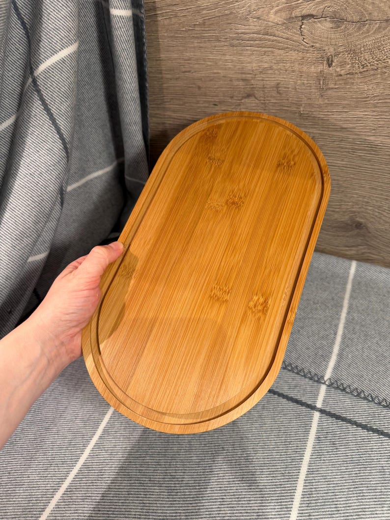 May include: An oval-shaped bamboo serving tray with a smooth, light brown wood grain finish. The tray features a raised outer rim and a flat, recessed center surface, showcasing the natural texture and color variations of the bamboo material.