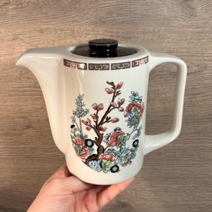 May include: White ceramic teapot with a black lid and a handle. The teapot is decorated with a floral design in shades of red, pink, blue, and green. The teapot has a spout for pouring.