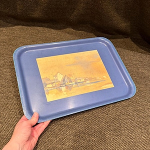 May include: A rectangular blue serving tray featuring a central rectangular illustration of a coastal harbour scene with buildings, boats, and water in muted yellow, brown, and blue tones. The tray has raised edges and a smooth, solid blue border.