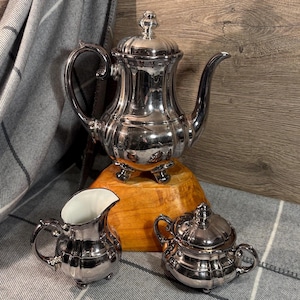 May include: A silver tea set with a teapot, creamer, and sugar bowl. The teapot has a curved spout and a decorative lid. The creamer and sugar bowl have small handles. The set is displayed on a wooden base and a gray blanket.