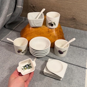 May include: A ceramic serving set featuring white square bowls, round cups with olive illustrations, small white spoons, and stacked white saucers. The collection includes a small square dish with a painted olive branch motif and several plain white plates.