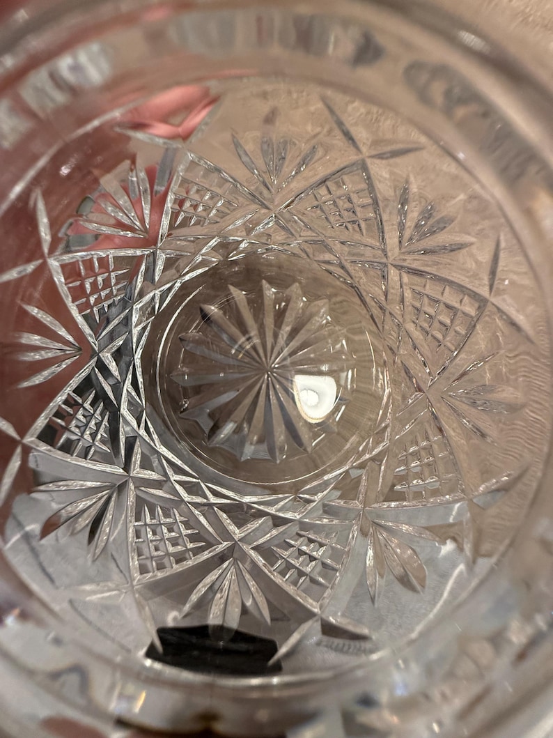 Op de afbeelding: Close-up van een helder glas met ingewikkelde geometrische patronen. Het ontwerp bevat stervormige en rasterachtige vormen, waardoor een textuur ontstaat. Het glas lijkt leeg, met licht dat weerkaatst op het geslepen kristal.